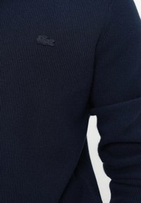Pulover texturat de culoare albastru marin cu un design PLIAT, având un logo brodat mic, închis, pe piept. Material moale și mâneci ajustate.
