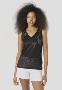 Fekete ujjatlan felső V-nyakkal, texturált anyagból, aprólékos gyöngydíszítéssel a vállrésznél. Fehér rövidnadrággal párosítva.