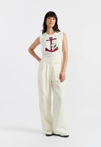 Cremfarbene, gestrickte ärmellose Bluse mit rotem Anker und Cartoon-Charakter-Design, kombiniert mit lockeren, hellbeigen Hosen und schwarzen Pumps.