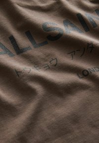 Ruda medvilninė marškinėliai su tamsiu tekstu, įskaitant "ALLSAINTS" ir japoniškus simbolius, su minkšta tekstūra ir kasdiene dizainu.