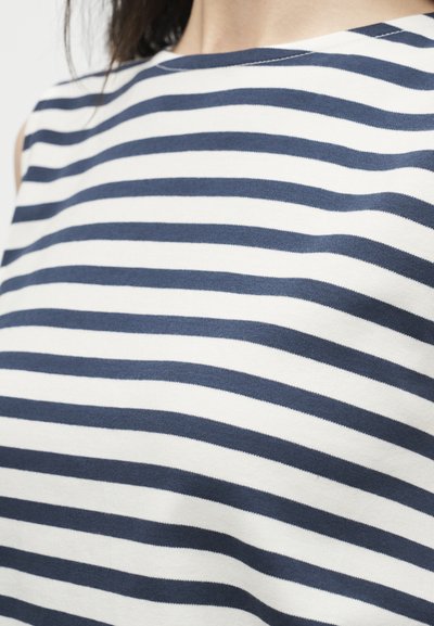 Γυναίκα που φοράει λευκό τοπ χωρίς μανίκια με οριζόντιες ναυτικές μπλε ρίγες, με εμφανές το λαιμό και μέρος του ώμου.