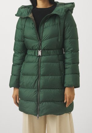 Femme portant un manteau matelassé vert foncé avec capuche, fermeture éclair à l'avant et ceinture assortie, associée à un pantalon beige et un pull noir.