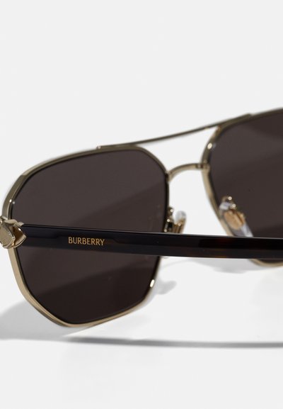 Óculos de sol com armação metálica dourada e lentes escuras. As hastes são em tartaruga escura com o logótipo "Burberry" em letras douradas.