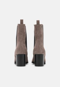 Une paire de bottines en daim taupe avec des panneaux stretch noirs sur les côtés et des talons bloc, vues de dos sur un fond blanc.