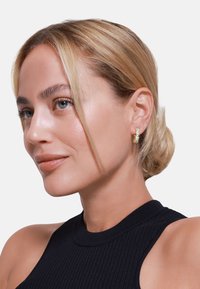 Zlaté kruhové náušnice s priehľadn�ými drahokamami, s vrstveným dizajnom. Model má rovné blond vlasy a nosí priliehavý čierny rebrovaný top.