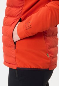 Giacca puffer rossa con tessuto testurizzato, dotata di una tasca con zipper e di una manica nera con testo stampato. Include un accento verde.