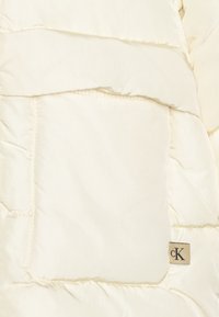 Balta pūkinė striukė su tekstūruotu audiniu, dideliu kvadratiniu kišeniu ir mažyte CK logotipo etikete kreminės spalvos.