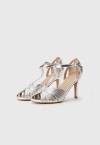 Silberne Sandalen mit offenem Zehenbereich, ausgestattet mit überkreuzten Riemen und einem schlanken Knöchelriemen. Glatte metallische Oberfläche und spitzes Design des Absatzes.