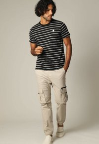 Schwarz-graue gestreifte kurzärmelige T-Shirt, kombiniert mit beigen Cargo-Hosen mit Reißverschlusstaschen und elastischen Bündchen. Weiße Slipper.