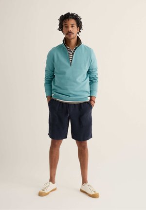 Muž stojaci s rukami v náprsnom vrecku námorníckych šortiek, oblečený v tyrkysovej mikine so štvrťzipsom, pruhovanej polokošeli, bielych teniskách a náramkoch.