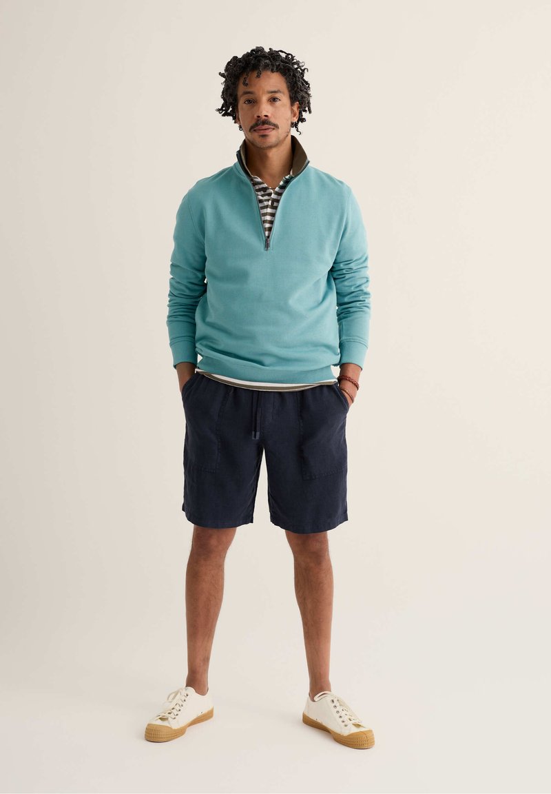 Homme debout les mains dans les poches de shorts bleu marine, portant un sweat-shirt à fermeture éclair courte bleu sarcelle, un polo rayé, des baskets blanches et des bracelets.