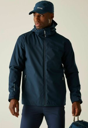 Man met navy waterdichte jas en pet, houdt blauwe rugzak vast aan riem, kijkt opzij tegen een neutrale achtergrond.