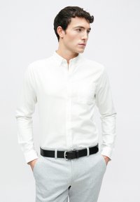 Cămașă albă cu nasturi, mâneci lungi, buzunar simplu la piept, asortată cu pantaloni gri deschis și curea din piele neagră.