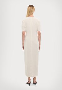 Robe longue couleur crème à manches courtes, silhouette droite, avec un détail de couture au dos, portée avec des sandales noires à talons.