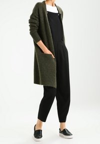 Long cardigan en maille olive sur une combinaison noire, avec des bretelles réglables, des poches latérales et des baskets noires à enfiler.