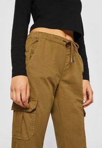 Pantalones cargo caqui con cintura elástica, acento de cordón, bolsillos laterales y un bolsillo con solapa en el muslo. Tejido texturizado, corte casual.