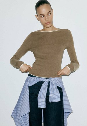 Jeune femme aux cheveux plaqués en arrière, portant un haut marron à manches longues, un jean foncé et une chemise rayée bleue nouée autour de la taille.