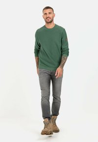 Green crew-neck sweater with ribbed texture, paired with light gray jeans and tan lace-up boots, walking on a white background.