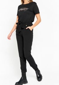 Vrouw in zwarte hoge taille broek met strikceintuur, zwart graphic t-shirt en zwarte stevige Chelsea boots, staand tegen een witte achtergrond.