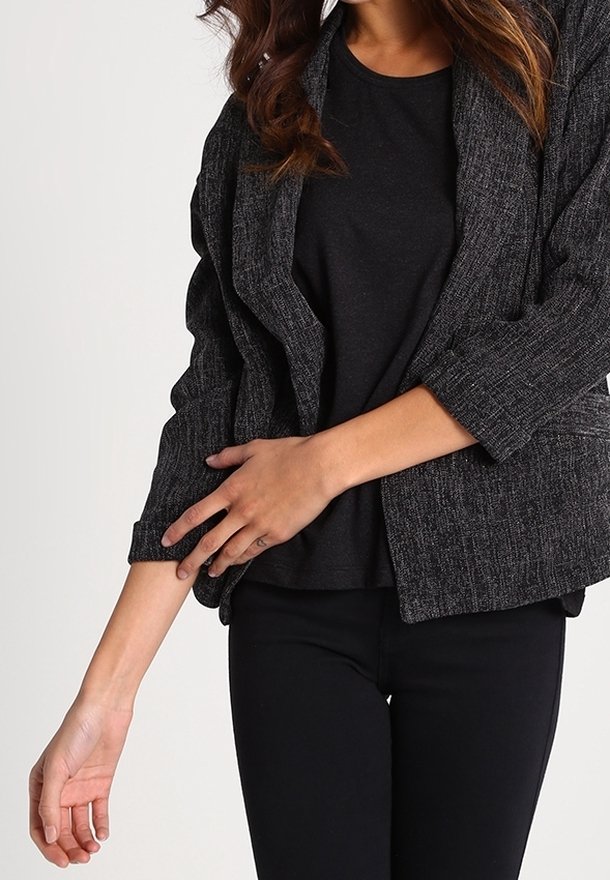 Femme ajustant la manche d'un blazer texturé foncé porté sur un haut noir et un pantalon noir, sur un fond clair uni.