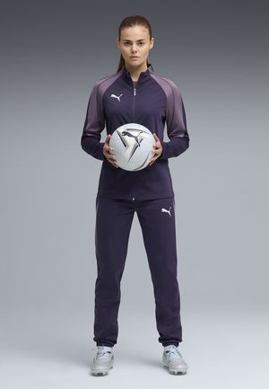 Kobieta w fioletowym dresie marki Puma, trzymająca białą piłkę nożną Puma, stojąca na tle szaro-gradientowego tła, zwrócona przodem.