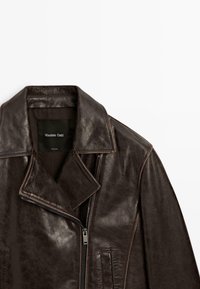 Braune Lederjacke mit weitem Kragen, Reißverschluss und zwei Seitentaschen. Weiches, strukturiertes Material mit dezentem Glanz. Markenetikett sichtbar.