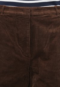 Detailný záber na opasok a rozopínanie hnedých kordurových nohavíc, nosených s námornícky modrým a bielym pruhovaným topom viditeľným v oblasti pása.