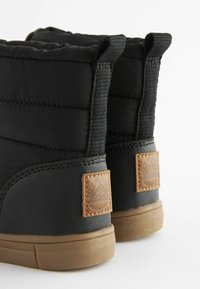 Bottes noires matelassées avec une tige rembourrée, des accents en cuir marron texturé et une semelle en caoutchouc. Présente une étiquette avec un texte sur le thème de l'aventure.