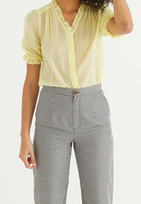 Blusa gialla trasparente con scollatura arricciata e maniche; chiusura con bottoni. Pantaloni a vita alta in gingham bianco e nero con pieghe.