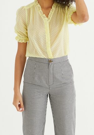 Blusa gialla trasparente con scollatura arricciata e maniche; chiusura con bottoni. Pantaloni a vita alta in gingham bianco e nero con pieghe.