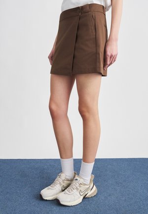 Młoda osoba ubrana w brązową spódnicę, białe skarpetki i beżowe sneakersy Nike stoi na niebieskim dywanie.