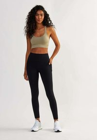Brassière de sport vert olive avec un design court, associée à un legging noir taille haute avec des poches latérales et un tissu texturé. Baskets blanches.