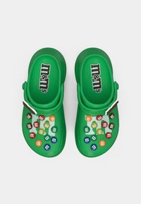 Green clogs with M&M's logo. Upper features colorful M&M's candies, textured surface, and a small adjustable strap.