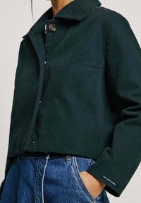 Ciemnozielona, krótka kurtka z wyprofilowanym kołnierzem, srebrnym zapięciem na guziki, kieszenią na klatce piersiowej oraz niebieskimi dżinsami z wykończeniem w postaci podwójnego szwu.