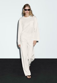 Witte longsleeve top met een zak, gecombineerd met losse witte broek en zwarte zonnebril; gladde stof, relaxed fit, minimalistisch design.