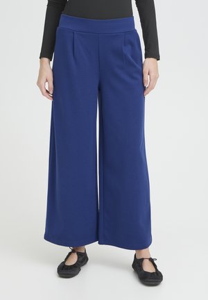 Persona che indossa pantaloni royal blu a gamba larga e vita alta con tasche laterali e ballerine nere, in piedi contro uno sfondo bianco.