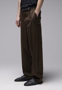 Pantalon marron à jambes larges avec une texture lisse. Comprend une ceinture noire et un détail chaîne. Associé à des mocassins noirs, porté par une personne.