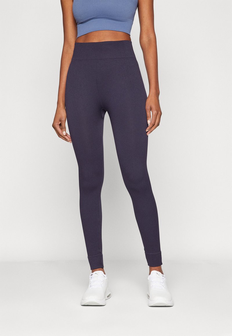 Ribbed dark purple leggings with a high waistband, tapered fit, and elastic material. Worn with white sneakers and a cropped blue top.
