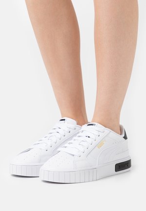 Baskets blanches Puma avec détail talon noir portées aux pieds sur un fond blanc uni, montrant les vues de côté et de face.