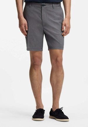 Man wearing gray tailored shorts and black lace-up shoes standing with hands relaxed by sides.