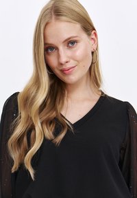 Schwarzes V-Ausschnitt-Top mit langen, durchsichtigen Ärmeln, die dezente Glitzereffekte und eine glatte Textur aufweisen. Das Model hat welliges Haar und trägt Ohrstecker.