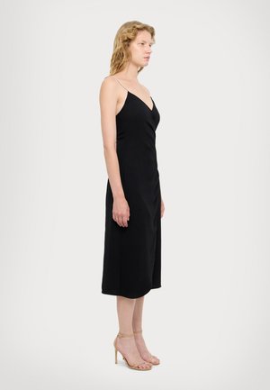 Abito nero di media lunghezza con scollatura a V e sottili spalline. Tessuto liscio con una leggera lucentezza. Modello abbinato a sandali con tacco a strappo color pelle.