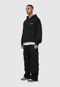 Svart oversized hettegenser med frontlomme og brodert logo. Kombinert med svarte cargo-bukser med flere lommer. Sneakers fullfører antrekket.