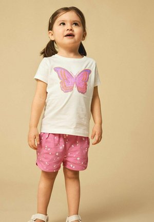 Weißes T-Shirt mit einem pinken und orangen Schmetterlingsdesign, kombiniert mit rosa Shorts, die kleine weiße Blumenmuster aufweisen. Heller Hintergrund.