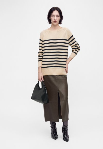 Femme portant un pull beige et noir rayé, jupe en cuir marron foncé avec une fente à l'avant, bottes noires, tenant un sac à main noir.