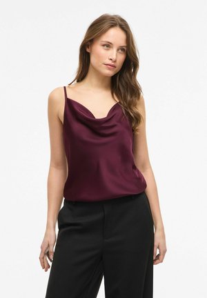 Femme aux longs cheveux bruns portant un haut en satin violet foncé sans manches et un pantalon noir, debout devant un fond blanc uni.