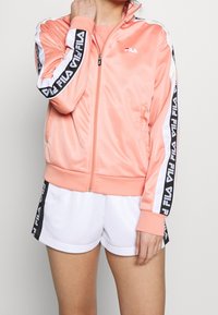 Pink satin zip-up jacket with white and black striped accents featuring Fila branding along the sleeves, paired with white shorts.