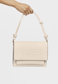 Borsa a spalla in finta pelle crema con logo "TOUS" impresso, forma piatta, manico superiore annodato e texture liscia.