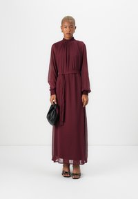 Pitkä burgundinvärinen mekko, jossa on korkea kaula-aukko, pitkät hihat ja rypytetty yksityiskohta. Mekossa on vyötärösolki ja se on tyylitelty mustilla asusteilla ja sandaaleilla.