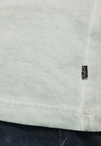 Lichtgrijs katoenen T-shirt met een rechte zoom, voorzien van een klein zwart label met witte stiksels aan de onderkant links.